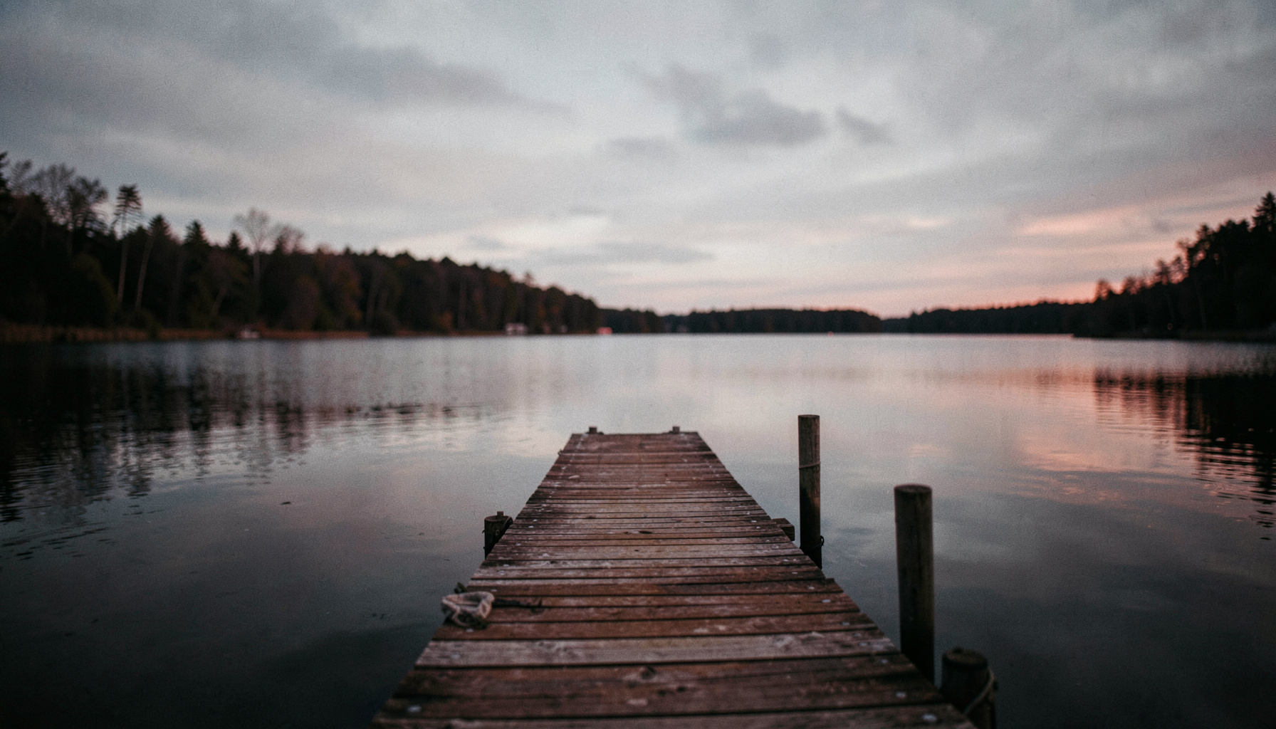 Wide cinematic view of a quiet lakeside dock at dusk, with muted rust and slate tones reflecting soft overcast light